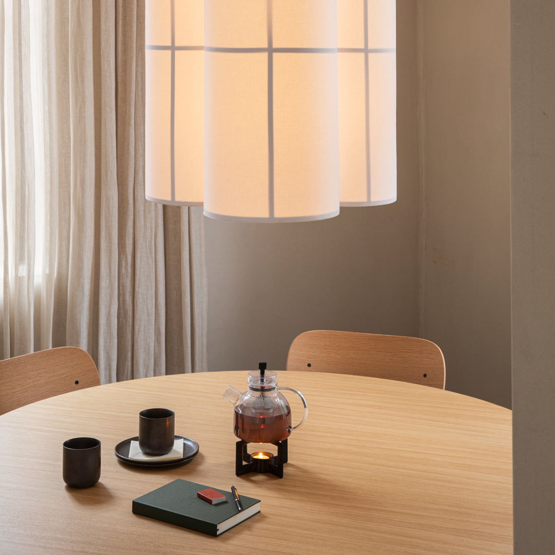 "Soft beige lamp above a round wooden table with black cups, a teapot, and a green book, creating a cozy atmosphere."
