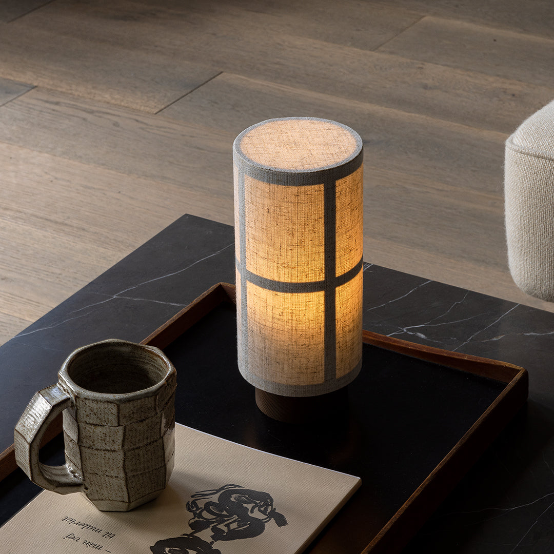 "Minimalist cylindrical lamp with a beige shade and gray accents, illuminating a warm glow on a dark wooden table."