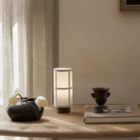 Minimalist lamp with a textured white shade and cylindrical design, set against a neutral-toned tabletop and backdrop.