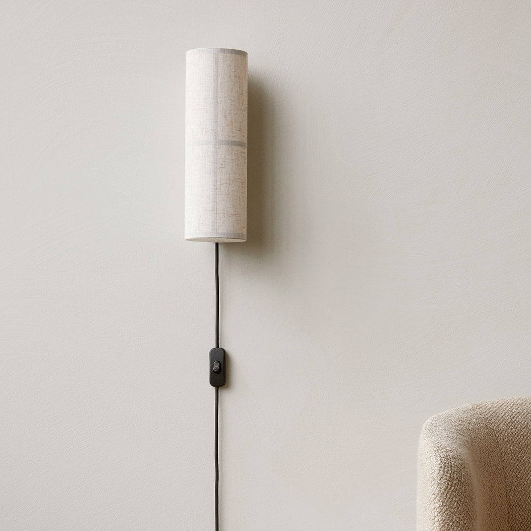 White fabric cylinder wall lamp with black cord and switch, minimalistic design against a neutral wall background.