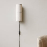 White fabric cylinder wall lamp with black cord and switch, minimalistic design against a neutral wall background.