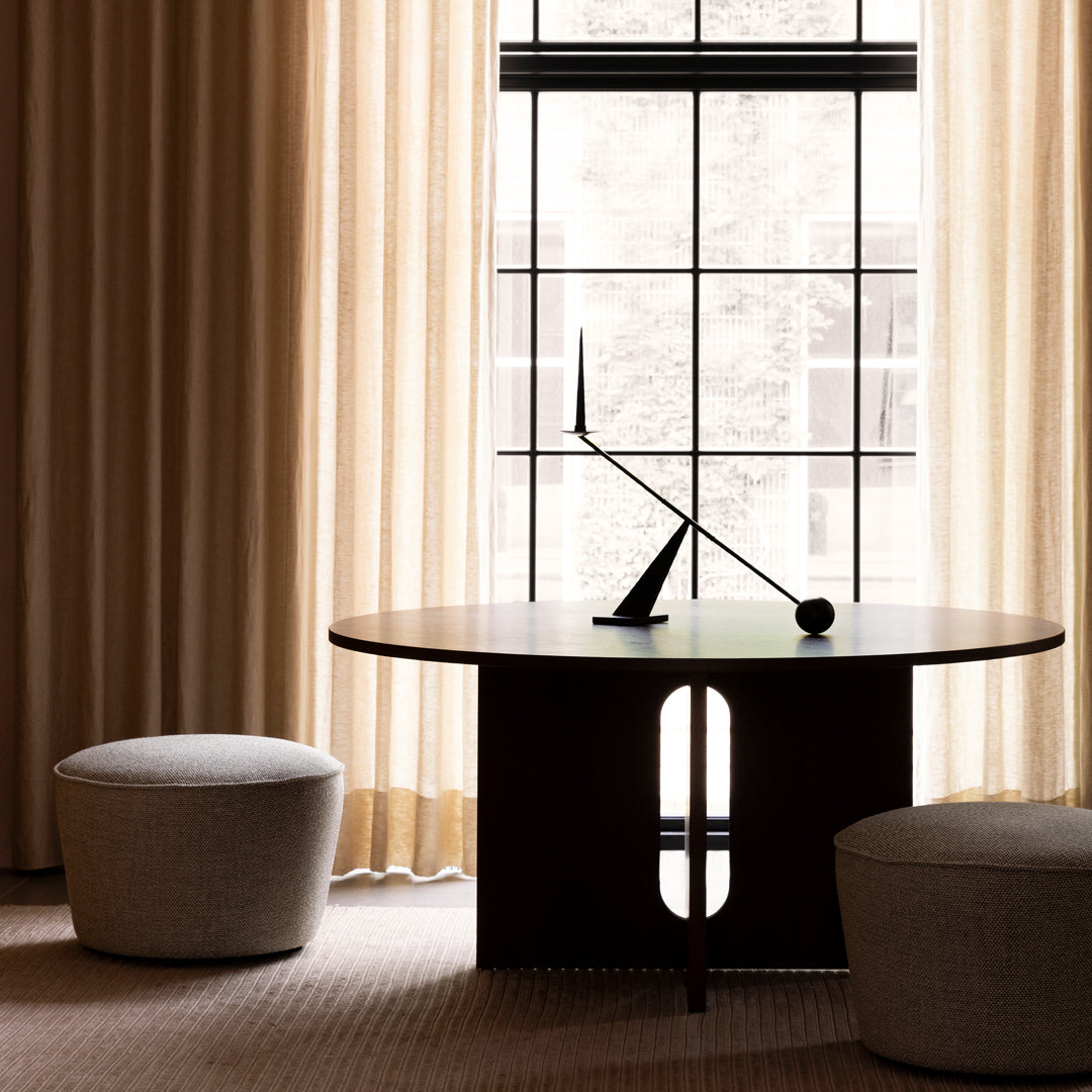 Modern round black table with sleek design, surrounded by soft beige poufs and warm, light-filtering curtains.