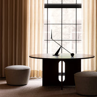 Modern round black table with sleek design, surrounded by soft beige poufs and warm, light-filtering curtains.