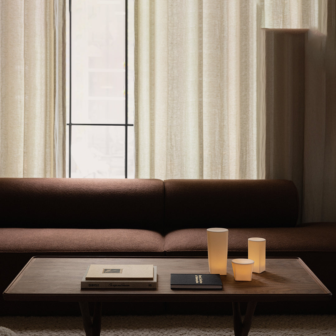 Brown sofa against textured light curtains, featuring soft beige candle holders and dark accent books on a wooden table.