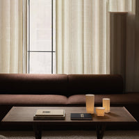 Brown sofa against textured light curtains, featuring soft beige candle holders and dark accent books on a wooden table.