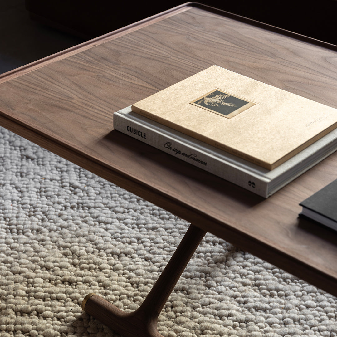 Brown wooden coffee table with a smooth surface, stacked books, and textured cream-colored rug below.