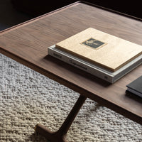 Brown wooden coffee table with a smooth surface, stacked books, and textured cream-colored rug below.