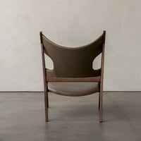 Brown upholstered chair with a curved design and cut-out accents, set against a neutral textured backdrop.