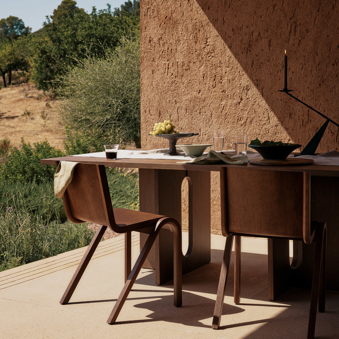 Modern wooden dining table in earthy tones with simple lines, paired with brown chairs against a warm, textured backdrop. 
