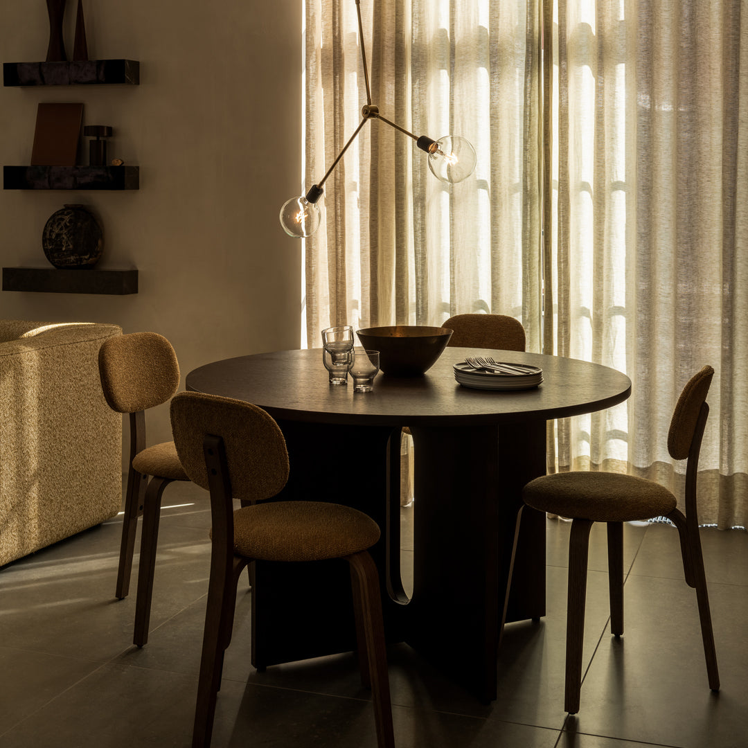 Modern round black dining table with tan upholstered chairs against soft beige curtains and a light-filled background.