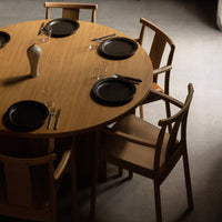 Round wooden dining table with natural grain, black plates, and elegant glassware set against a soft gray background.