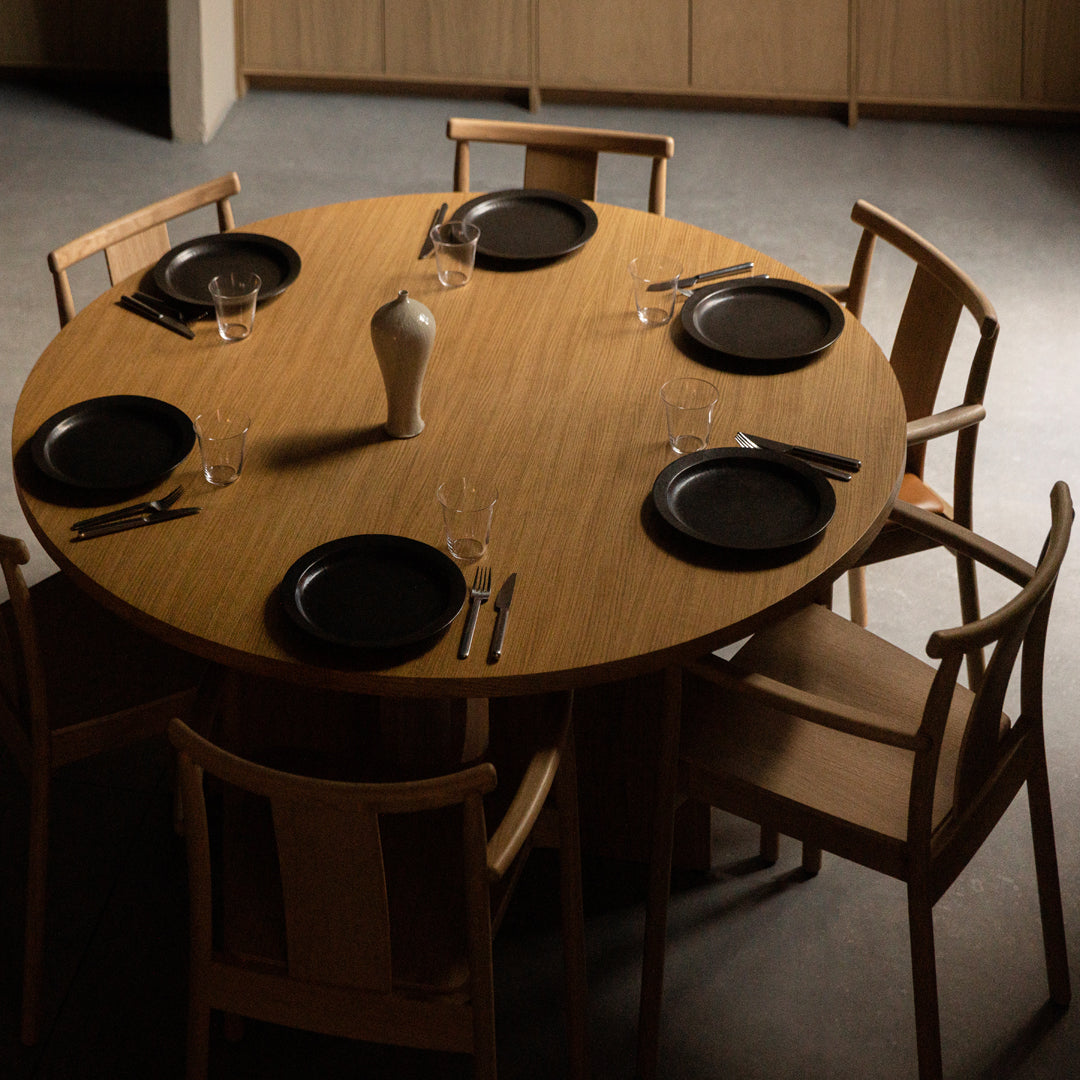 Round wooden dining table with six light wood chairs, featuring black plates and a beige vase centerpiece.