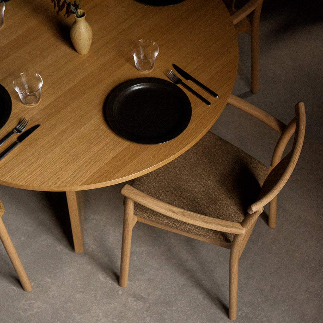 Round wooden dining table with natural oak finish, black plates, and neutral-toned upholstered chairs. Elegant, modern style.