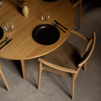 Round wooden dining table with natural oak finish, black plates, and neutral-toned upholstered chairs. Elegant, modern style.