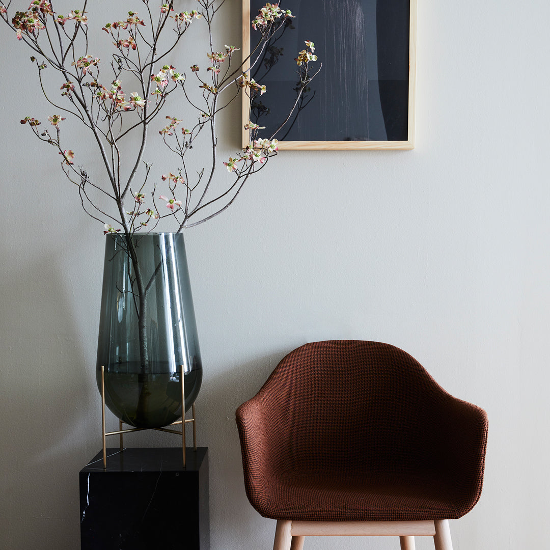 Brown upholstered chair beside a sleek black pedestal with a green glass vase and floral accents against a neutral backdrop.