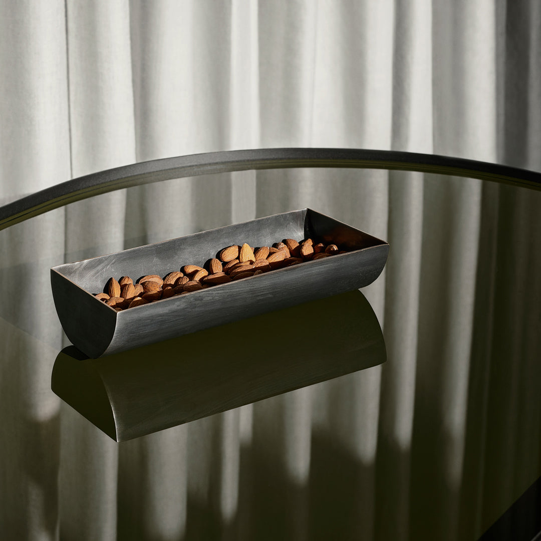 "Metallic gray bowl filled with brown almonds, reflecting on a dark glass surface with soft gray curtains in the background."