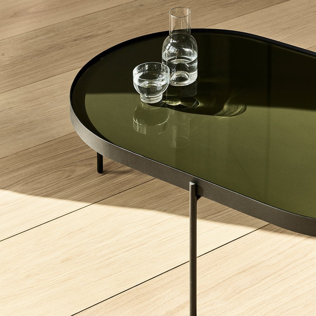 Oval coffee table with a dark green glass top and sleek black metal legs, set against a light wood floor.
