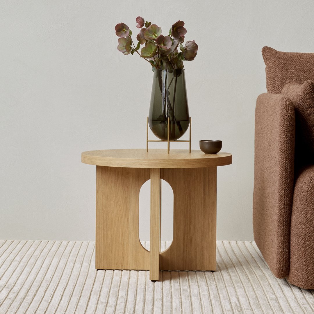 Natural wood side table with a circular top and cut-out design, paired with a muted brown sofa and a green vase.