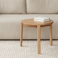 Light wood round side table on a textured beige rug, paired with a soft beige sofa. Simple, modern design.