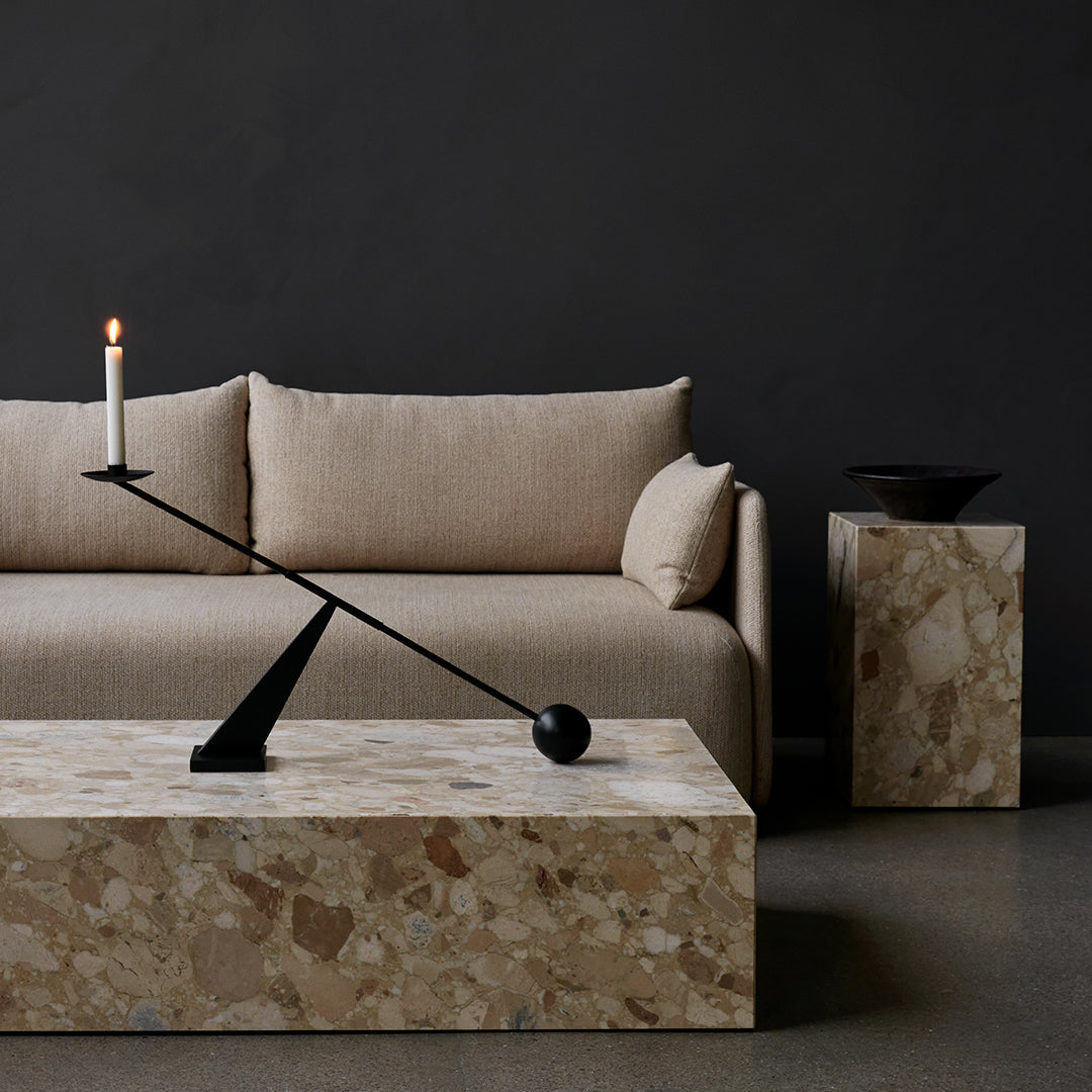 Beige linen sofa with a light brown stone table and black metal candle holder on a dark gray background.