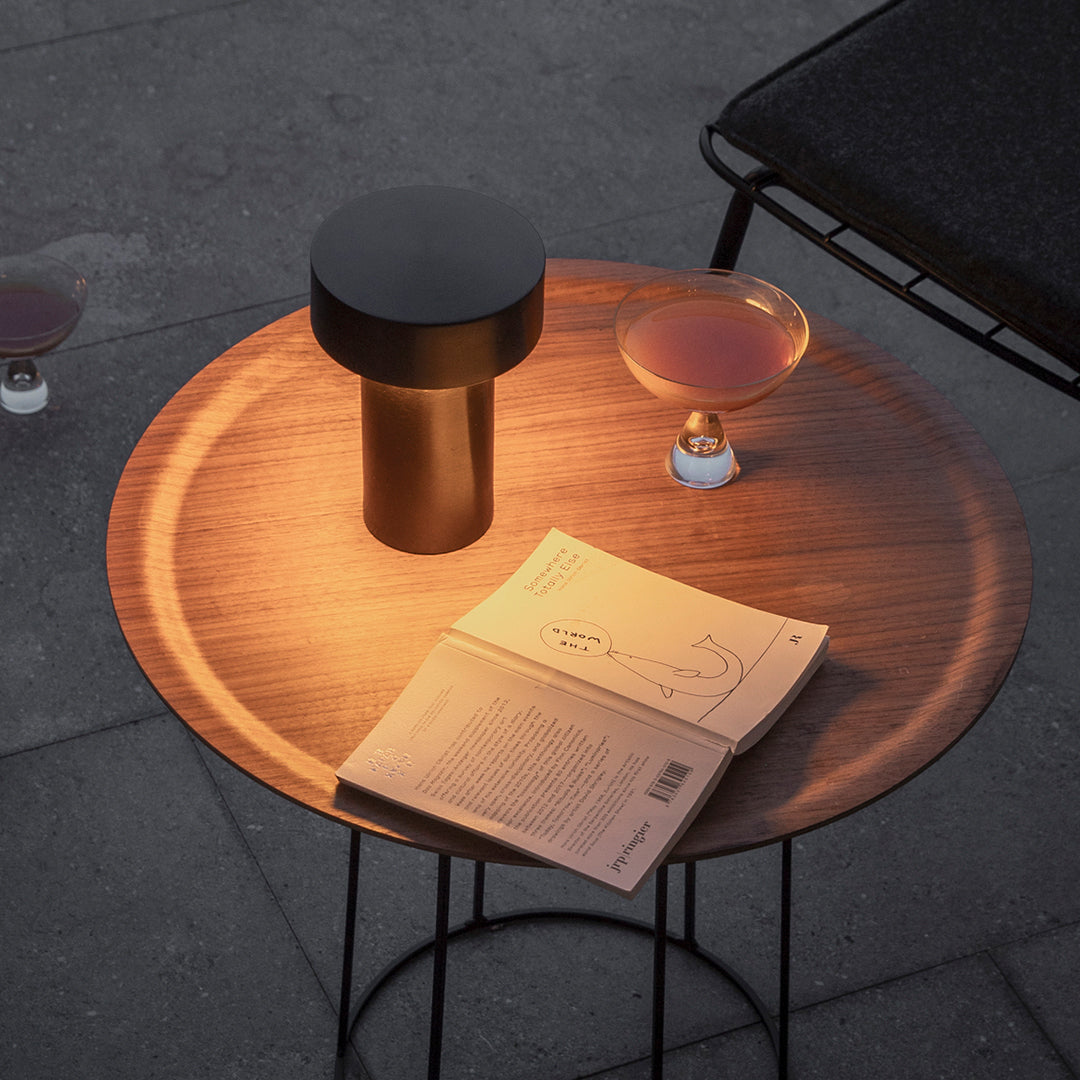Wooden round table with a sleek black lamp, clear cocktail glass, and open book on a textured surface.
