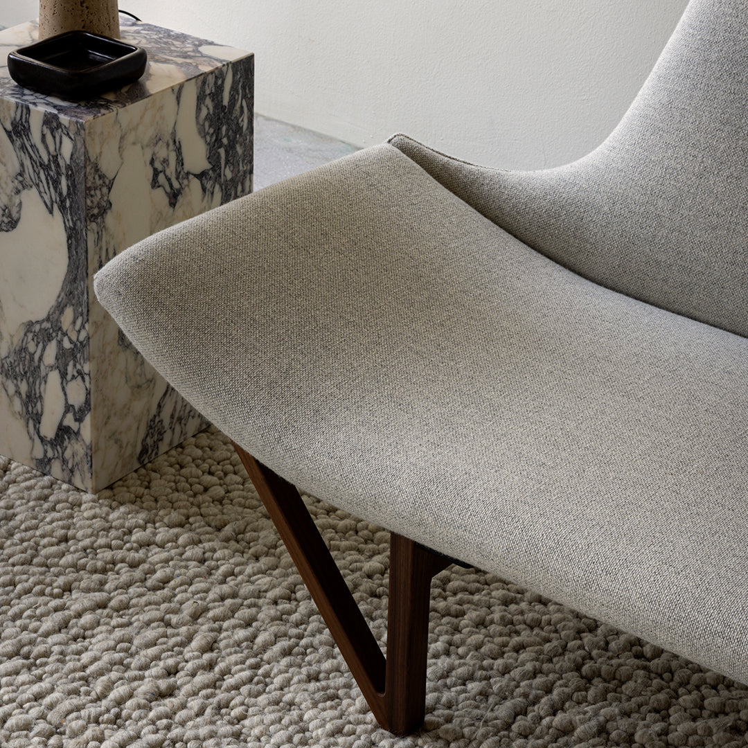 Grey upholstered chair with a smooth texture, featuring a wooden base and textured beige rug. Modern aesthetic.