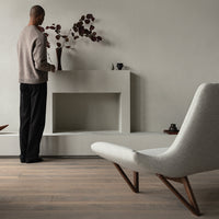 Neutral beige sweater, black pants, gray chair, earthy wooden floor, subtle vase with deep red leaves, modern minimalist backdrop.