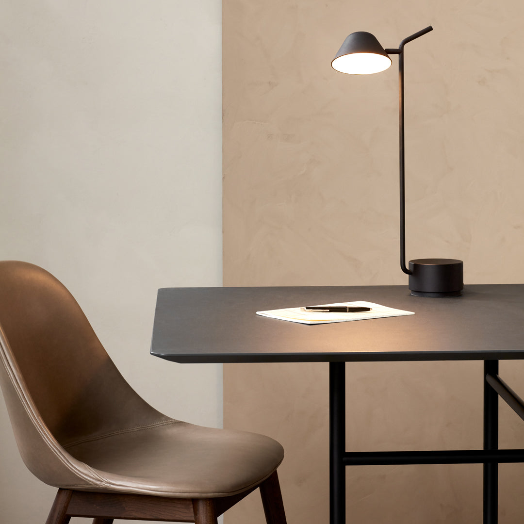 "Modern desk lamp with sleek black finish, paired with a warm brown chair against a minimalist beige wall."