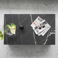 Black marble tabletop with white veining, accented by green leaves, black decor, and scattered newspapers.