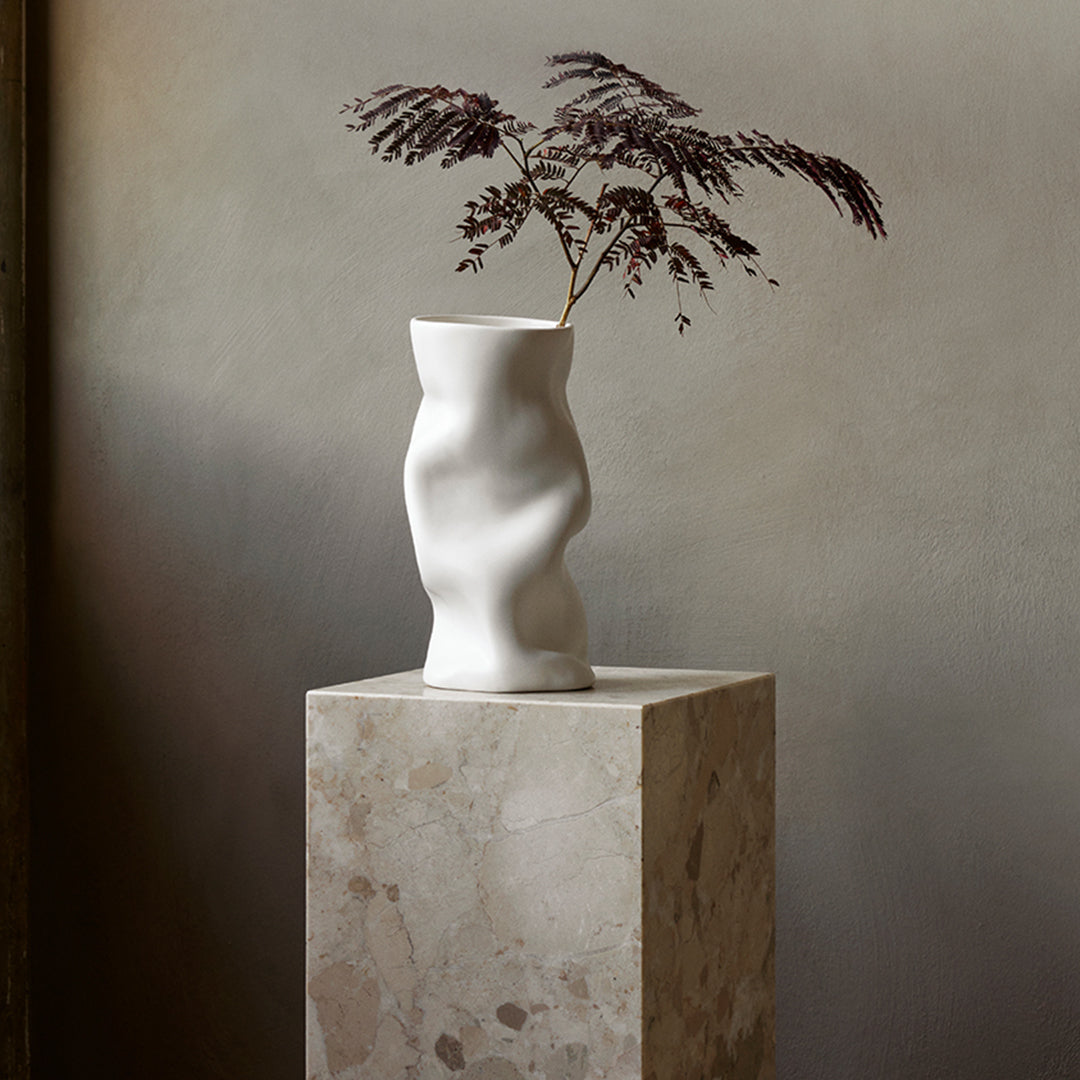 White abstract vase with a wavy texture and dark purple foliage on a natural stone pedestal against a neutral background.