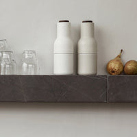White and light gray ceramic bottles with dark brown lids on a dark marble shelf, complemented by transparent glassware.