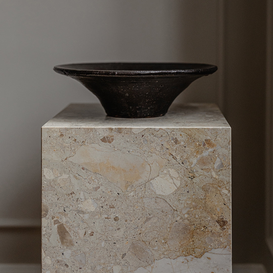 Black ceramic bowl on a textured beige stone pedestal, showcasing earthy tones and smooth, subtle patterns.