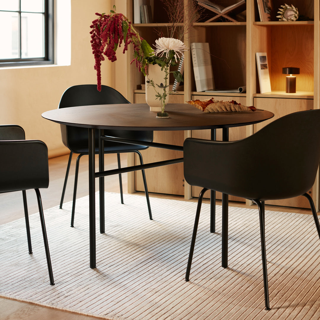 Round wooden dining table with black chairs on a textured cream rug, featuring natural decor and neutral tones.