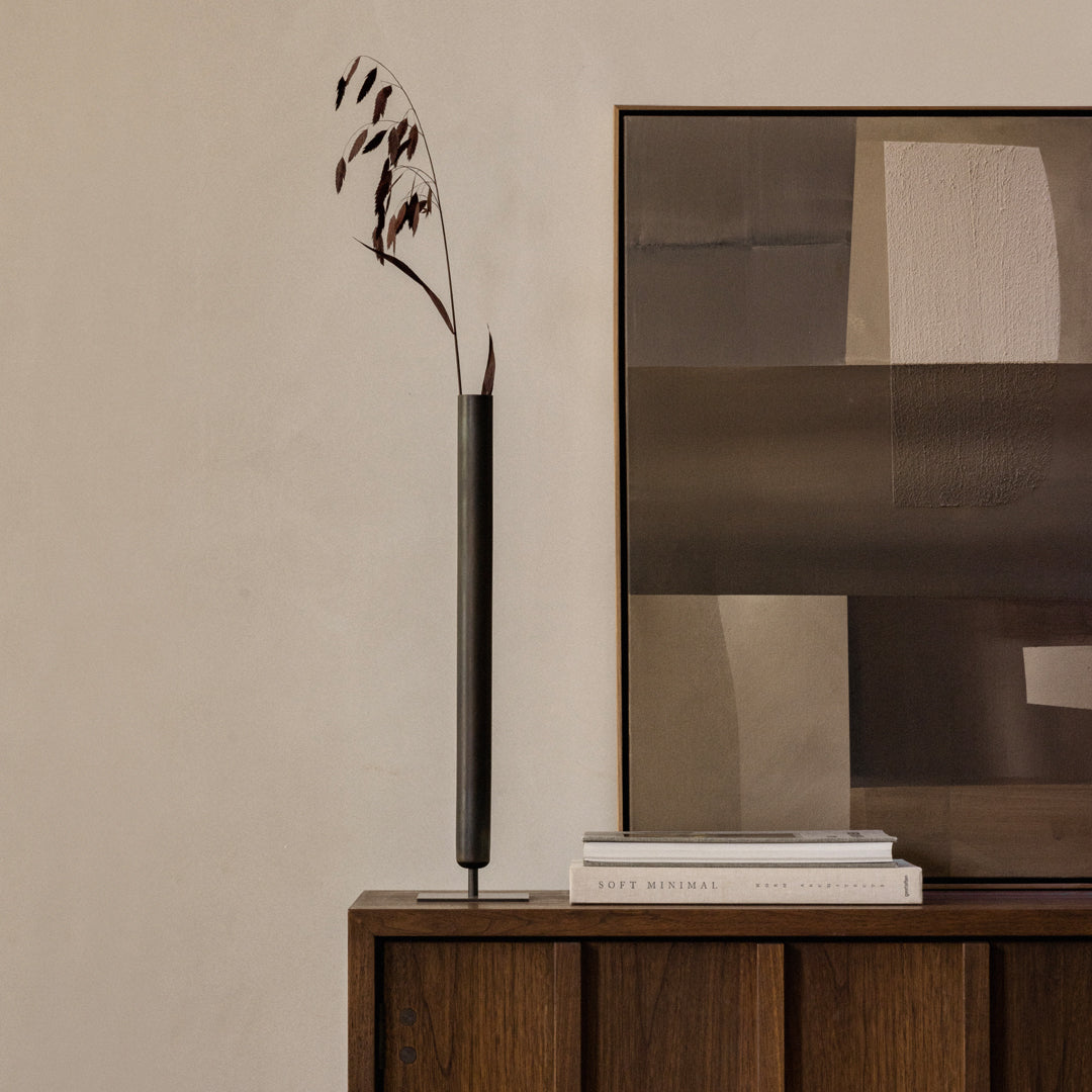 Black minimalist vase with dark brown foliage against a neutral textured wall and abstract art in muted tones.