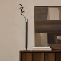 Black minimalist vase with dark brown foliage against a neutral textured wall and abstract art in muted tones.