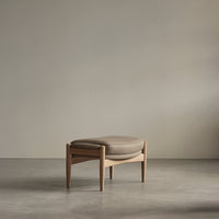 Beige upholstered ottoman with a minimalist wooden frame against a muted gray background. Modern and stylish design.