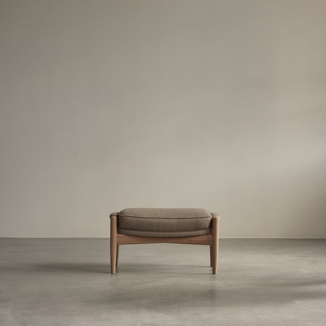 Brown upholstered ottoman with a minimalist wooden frame, set against a soft gray backdrop. Smooth, modern design.