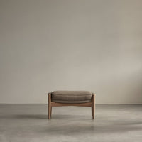 Brown upholstered ottoman with a minimalist wooden frame, set against a soft gray backdrop. Smooth, modern design.