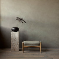 Minimalist gray wall backdrop with earthy tones, beige stone pedestal, and textured gray cushion. Elegant black vase accent.