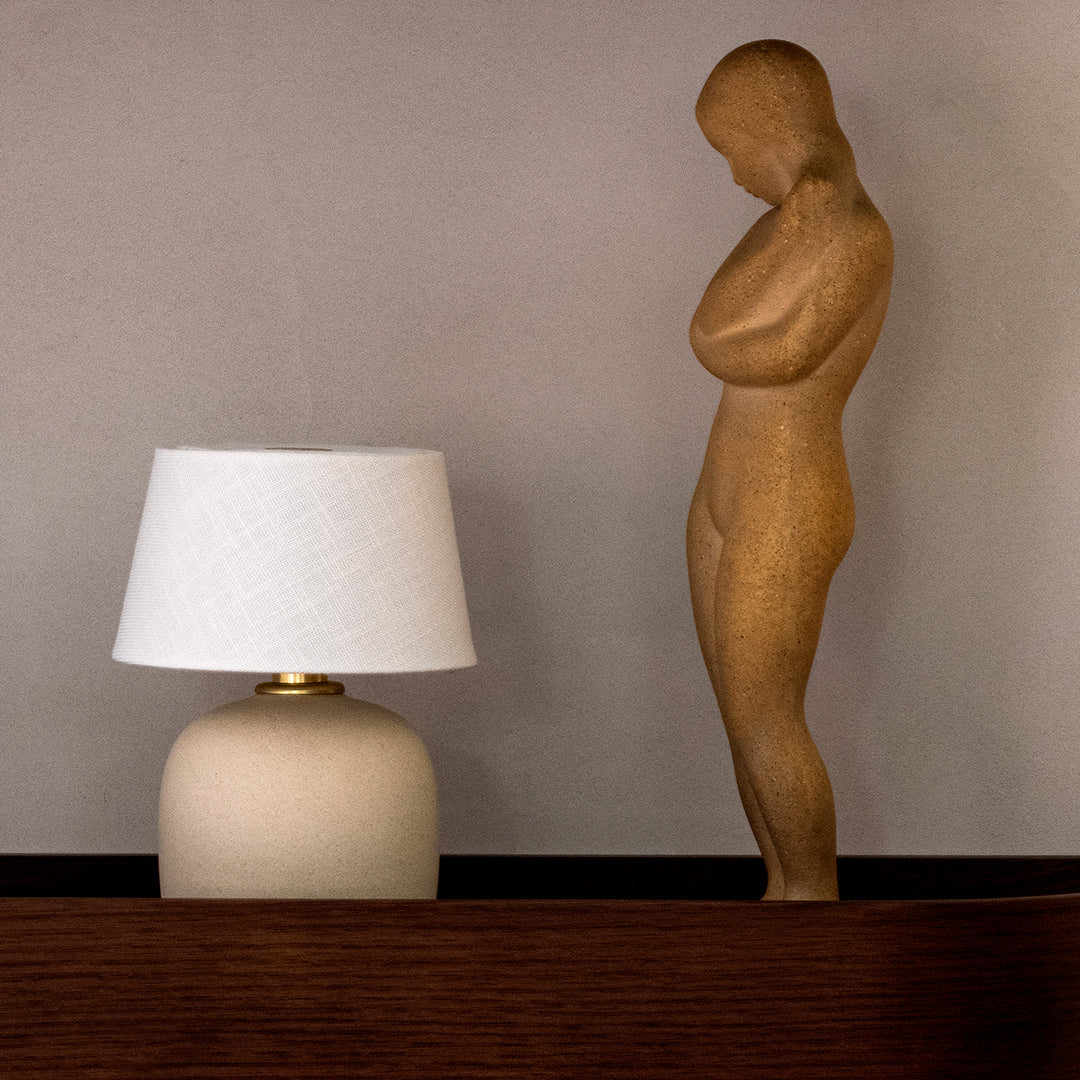 Neutral beige lamp with a textured shade beside a smooth, tan-toned wooden statue against a subtle gray backdrop.