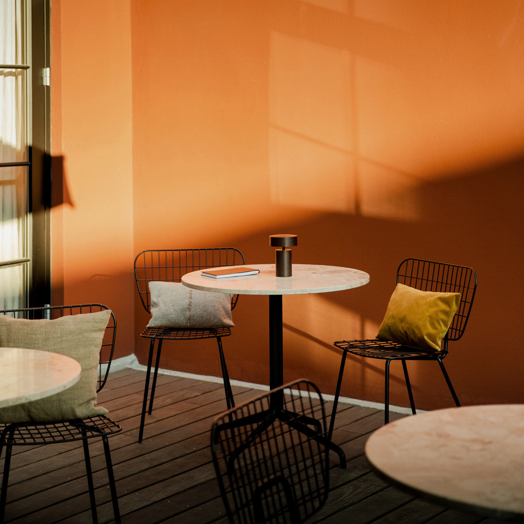 Modern outdoor seating with black wire chairs, marble tables, and yellow and beige cushions against an orange wall.