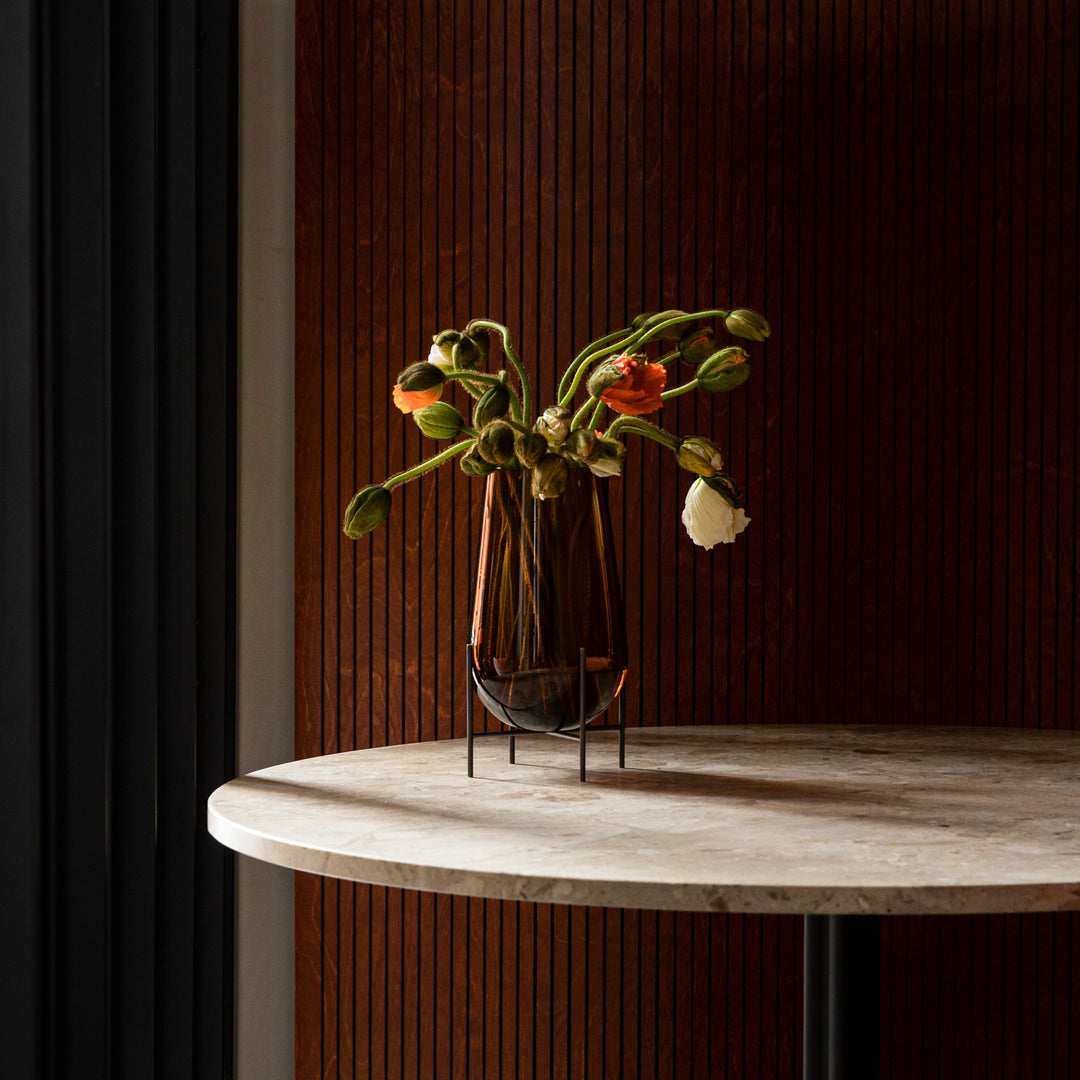 Amber glass vase with green and orange floral arrangement against a textured brown backdrop. Elegant and modern design.
