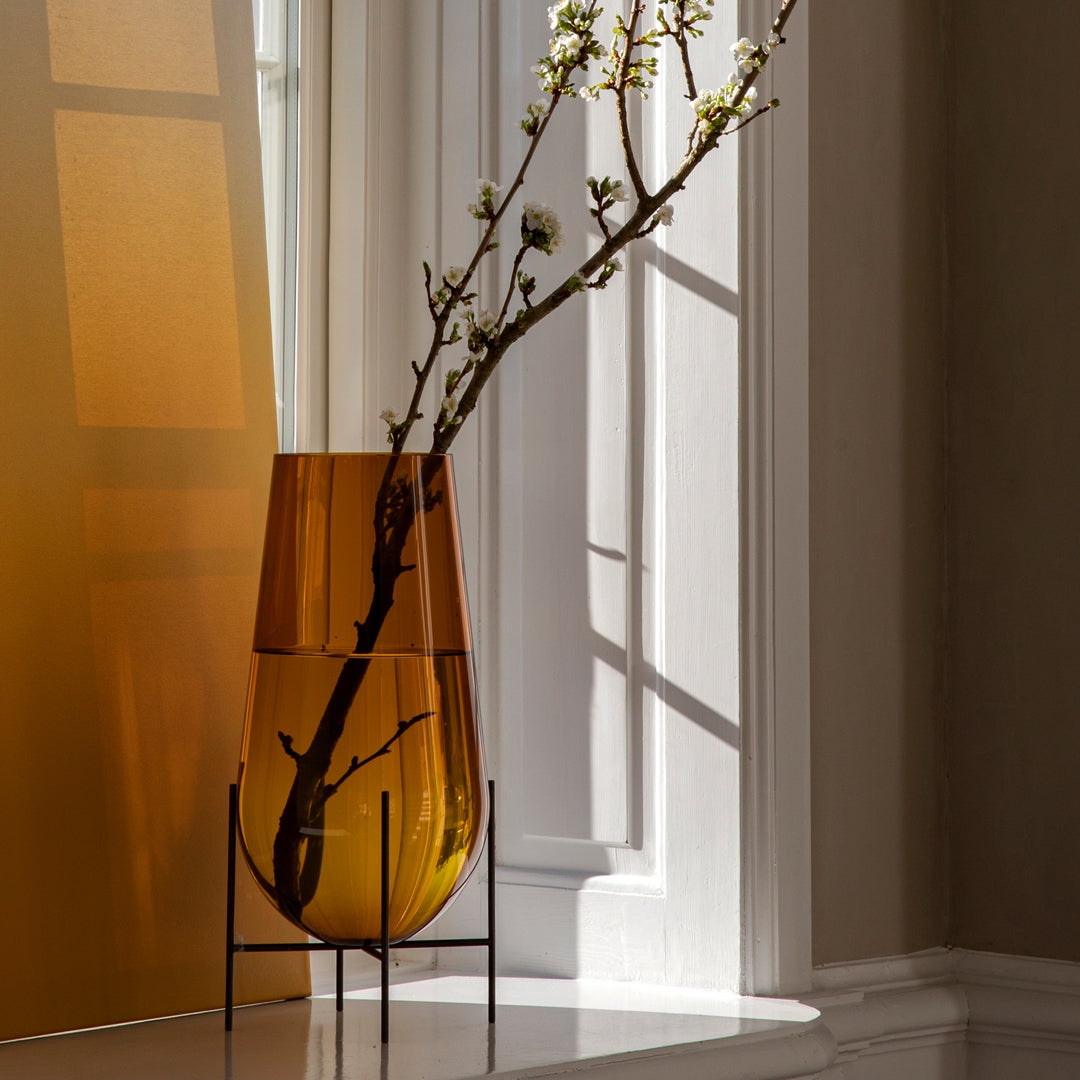 Amber glass vase with modern silhouette, featuring a branch, set against a warm, sunlit geometric background.