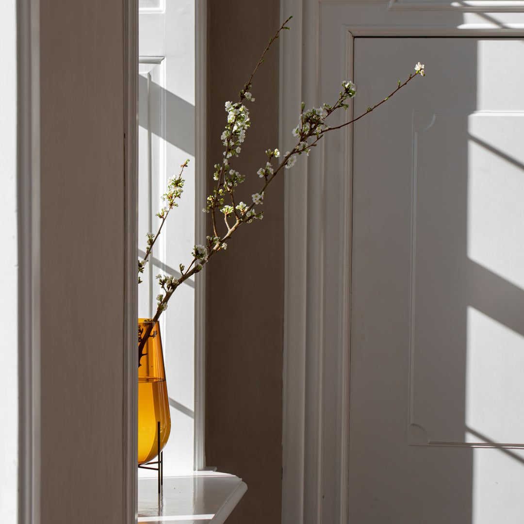 Bright amber vase with delicate branches in soft, natural light and contrasting white walls. Modern minimalist decor.