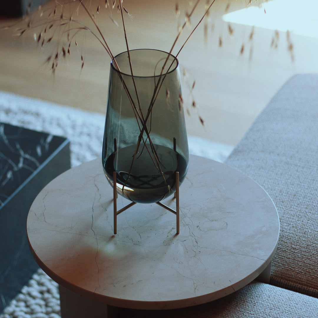 Smoky green glass vase in a minimalist brass stand, placed on a round white marble table with soft beige upholstery.