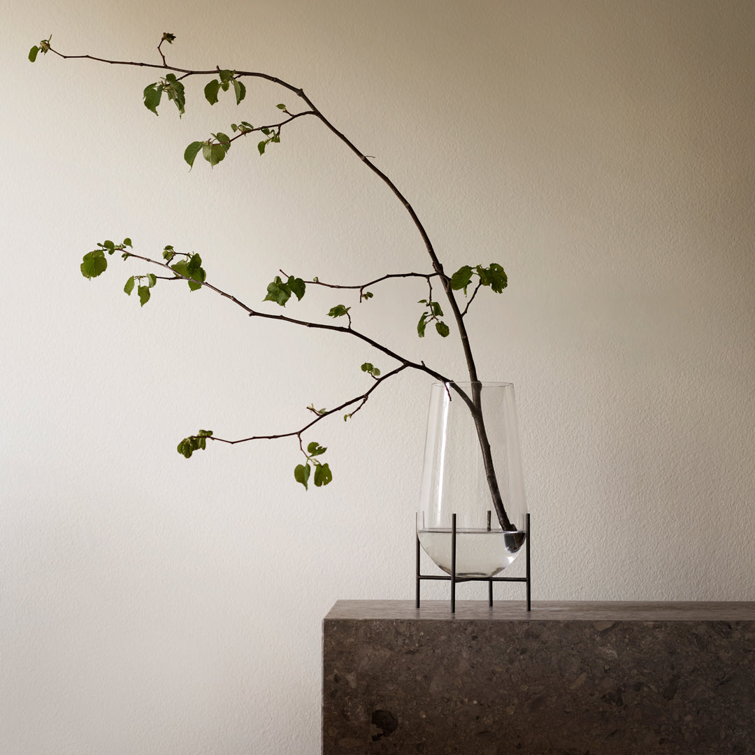Clear glass vase with slender black metal stand, showcasing vibrant green leaves against a neutral backdrop.