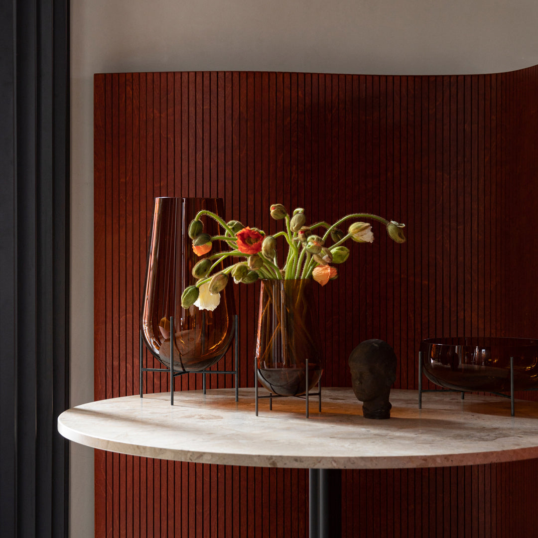 "Brown glass vases with floral arrangements on a round marble table, set against a textured, deep red backdrop."