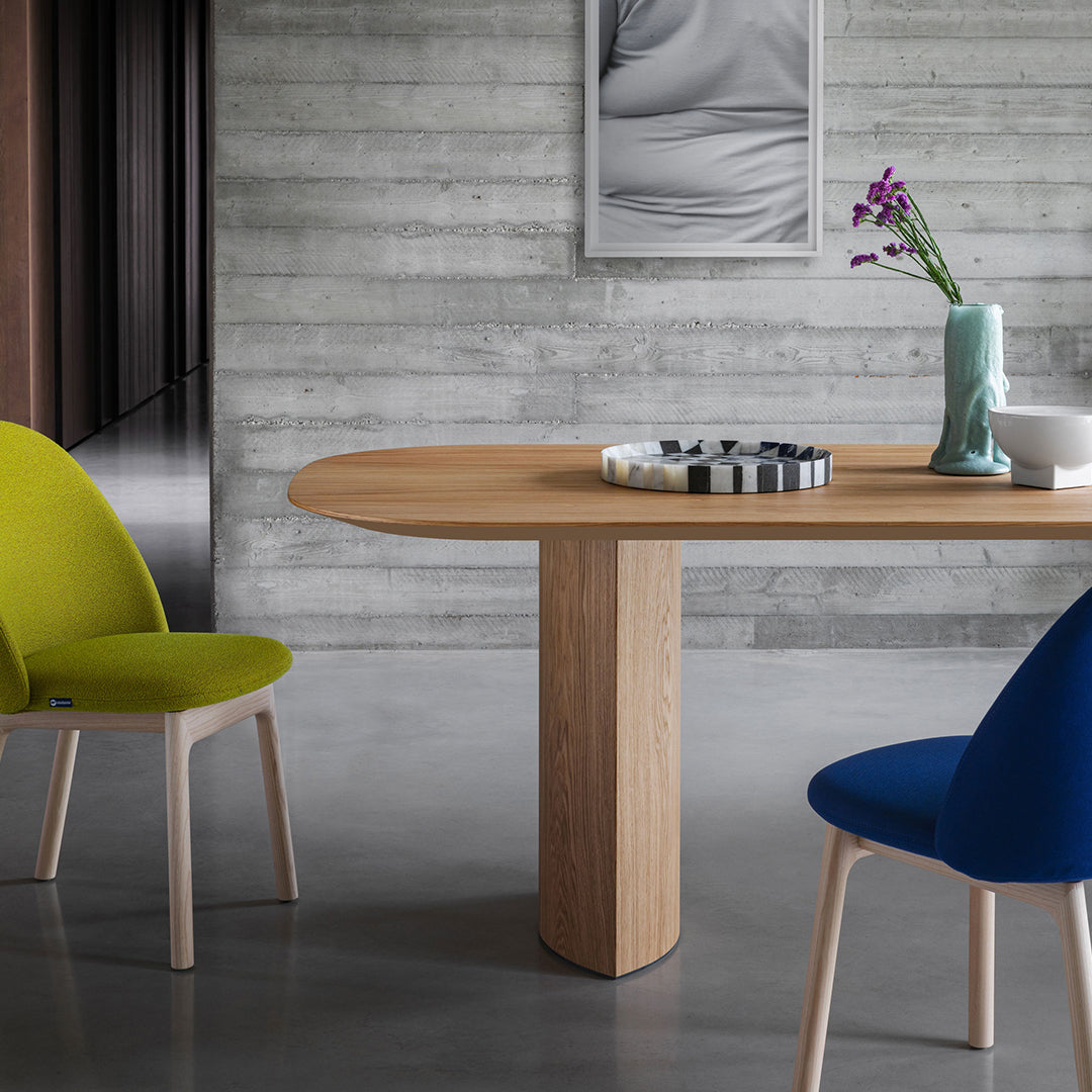 Modern wooden dining table with rounded edges, paired with vibrant green and blue chairs against a gray background.