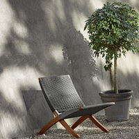 Stylish gray woven lounge chair with natural wood accents, set against a textured gray wall and greenery.