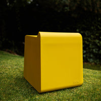 Bright yellow square ottoman with a smooth, modern finish, set against a green grass background.